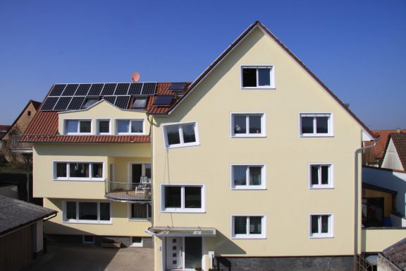 Gelbes Mehrfamilienhaus mit Solarpanelen und mehreren Fenstern unter klarem Himmel.